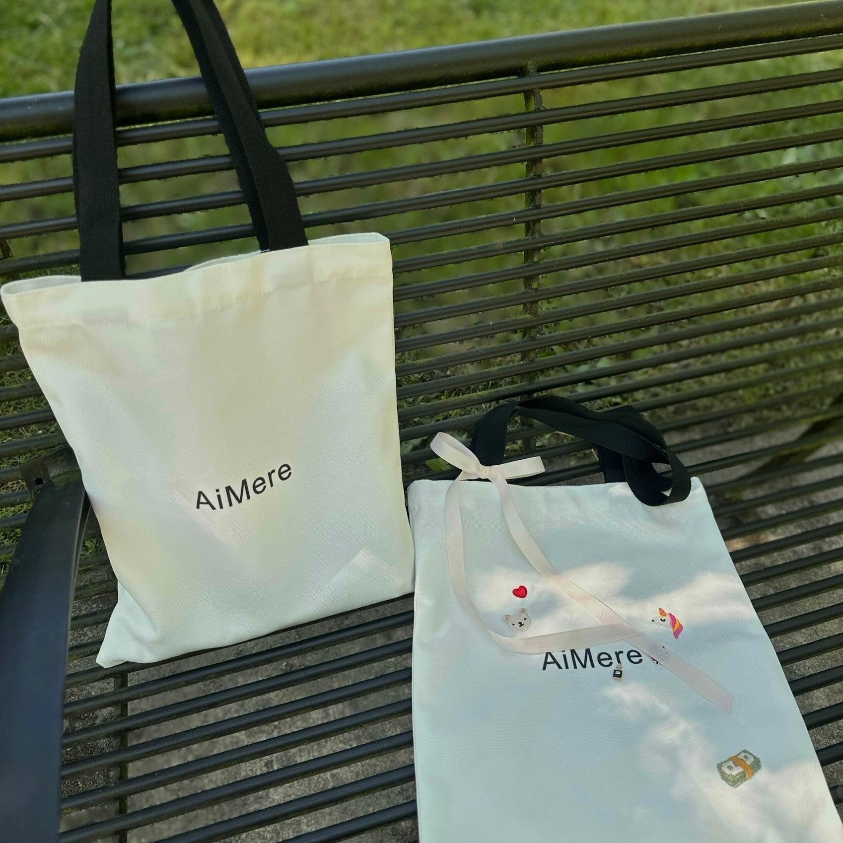 Two tote bags with 'AiMere' branding on a metal bench outdoors.