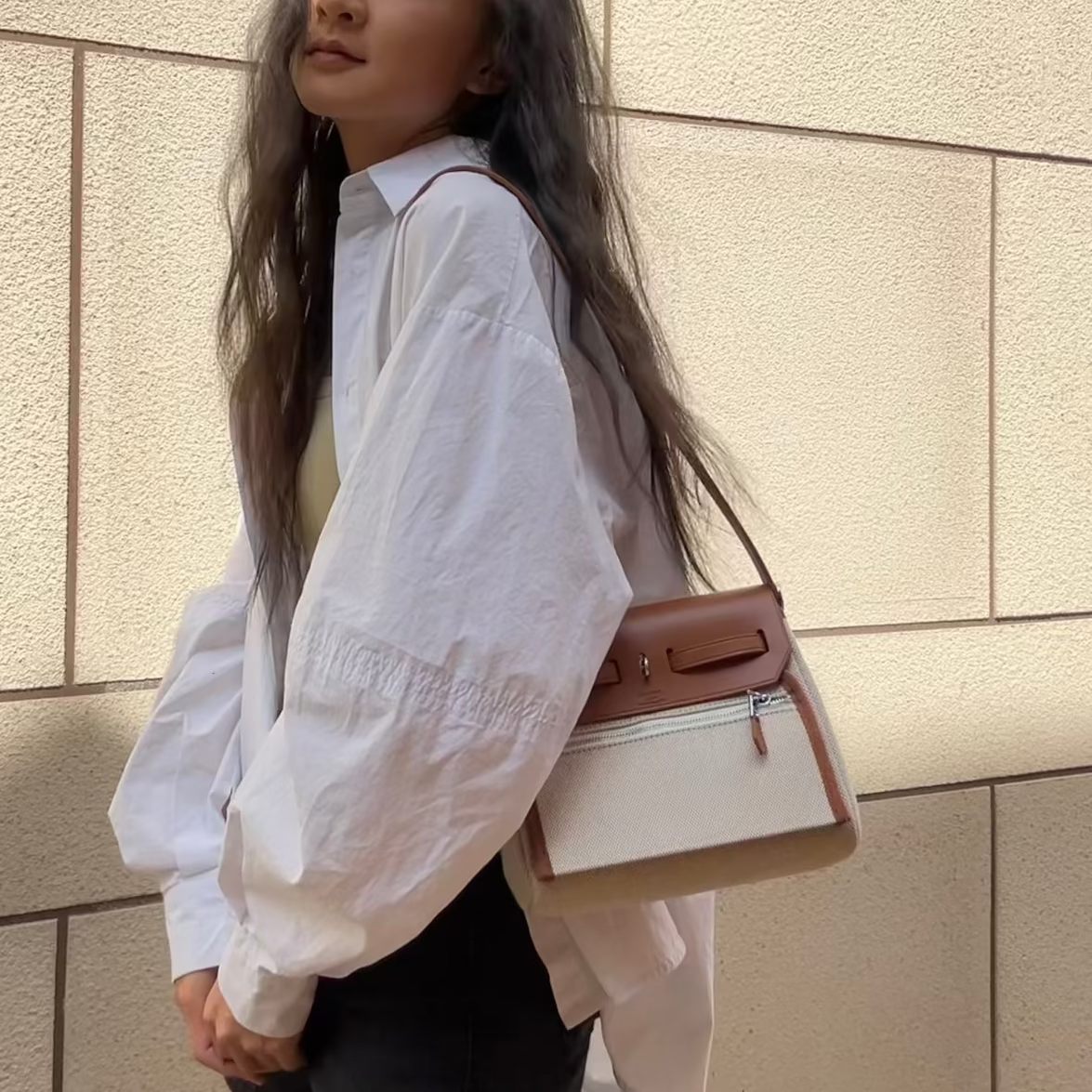 Woman in a white shirt and black skirt holding a gold Hermes bag against a beige wall.