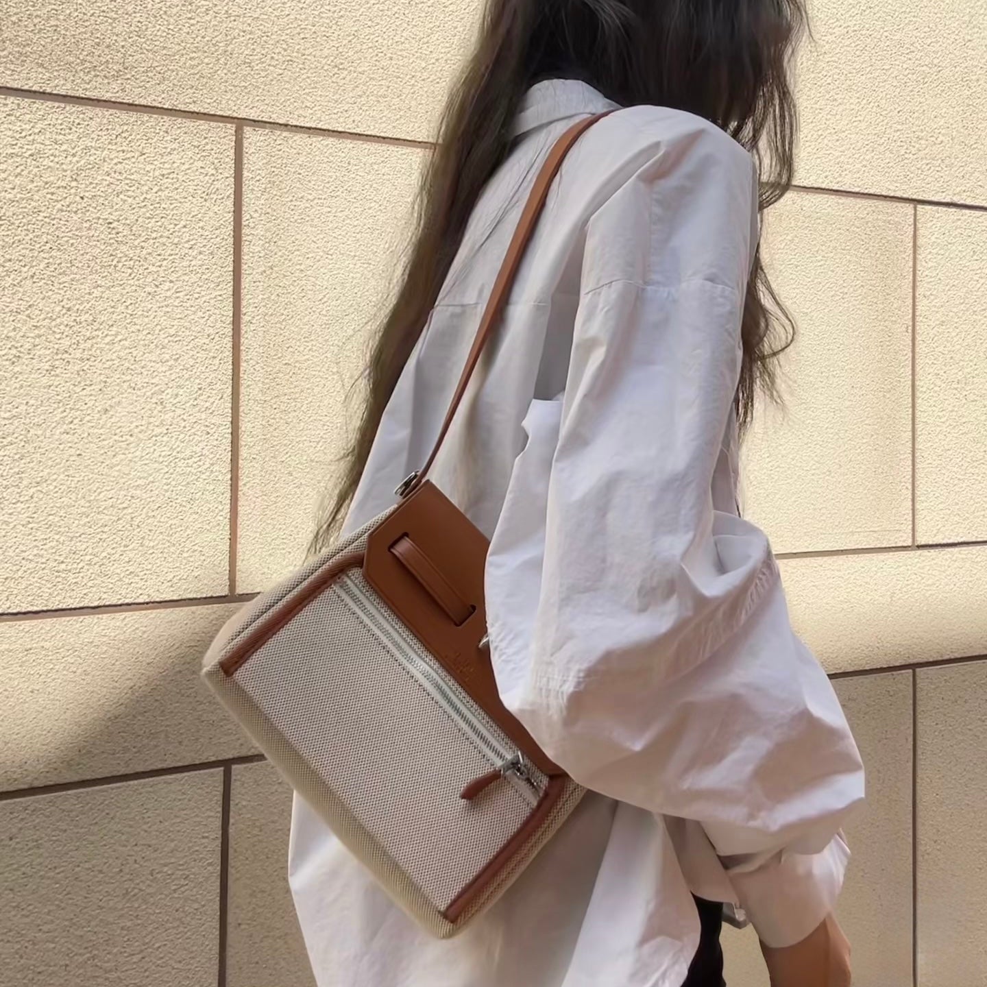 Person wearing a white shirt and carrying a beige and brown handbag against a beige wall.